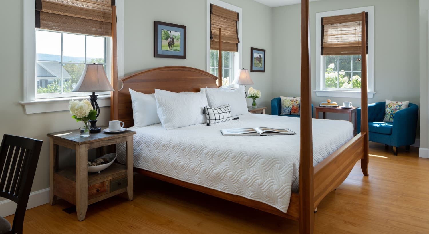 Bedroom with light colored walls, hardwood flooring, wooden four-poster bed, white bedding, wooden nightstands with lamps, and sitting area